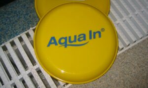 Eine gelbe Frisbeescheibe mit dem Aufdruck des AQUA IN Logos liegt unmittelbar am Beckenrand eines Schwimmbades. Das Bild dient als Blogbeitragsbild für den Blog, "Warum ist Sport treiben im Wasser gesund?"