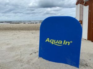 Im Bildvordergrung steht ein blaues Schwimmbrett mit der Aufschrift "Aqua In" in gelber Schrift vor einem Strandkorb. Zu sehen ist ein Sandstrand und in der Ferne Meer. Das Bild dient als Blogbeitragsbikd für den Blogartikel. "Wo mache ich Aquafitness?"