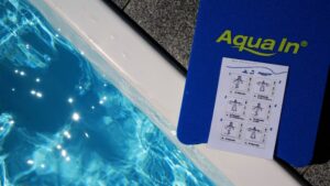 Ein blaues Schwimmbrett mit dem Aufdruck Aqua In liegt am Beckenrand eines Pools. Auf dem Brett liegt eine Bildanleitung für ein Aqua In Aquafitness Programm.