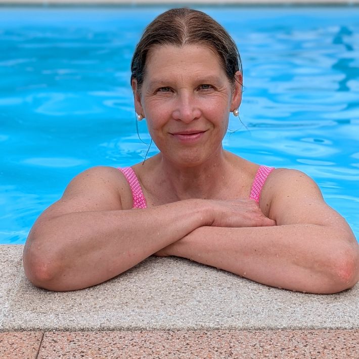 Portrait von Dr. Birgit Schmid, Gründerin von Aqua In, im Schwimmbecken. Sie steht im Wasser, lehnt mit verschränkten Armen am Beckenrand. Im Hintergrund schimmert blaues Wasser.