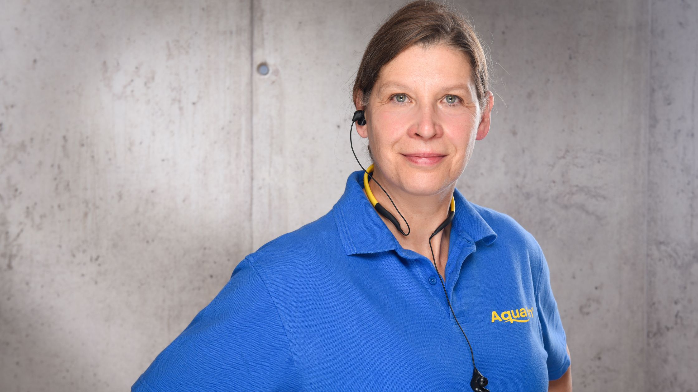 Portraitfoto von Dr. Birgit Schmid, in blauem T-Shirt vor einer grauen Wand. Um den Hals hat sie ein gelbes Headset.