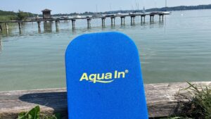 Ein blaues Schwimmbrett mit der Aufschrift "Aqua In" in gelber Schrift steht gegen ein Holzbrett gelehnt. Im Hintergrund ist ein See mit Segelschiffen zu sehen. Das Bild dient als Beitragsfoto für den Blogartikel. "Aquafitness für Anfänger - dein Einstieg darf leicht sein".