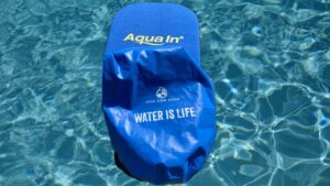Ein blauer Wassersack mit einer weißen Aufschrift: "VIVA CON AGUA - WATER IS LIFE" und ein blaues Brett mit gelber Aufschrift "Aqua In" liegen auf bewegtem blauen Wasser. Das Bild dient als Vorschaubild für einen Blogbeitrag.