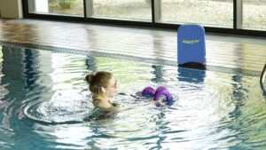 Das Bild zeigt eine junge Frau im Hallenbad, die Aquafitness betreibt. Sie trägt ein weißes Headset und übt mit lila farbenen Handgeräten, am Beckenrand steht ein blaues Brett mit der Aufschrift "Aqua In". Das Bild dient als Beitragsfoto für den Blogartikel "Aquafitness neu entdecken".