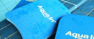 Blaue Schwimmbretter mit dem Logo AQUA IN liegen am Beckenrand. Dieses Bild dient als Beitragsfoto für den Blogbeitrag, "Wie gelingt Prävention in öffentlichen Bädern?"